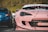 A close-up of a modified pink sports car with visible decals and custom bodywork. The focus is on the front end, showing intricate details such as a unique headlight design and body modifications. In the background, a blue car and a blurred figure are partially visible.