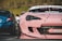 A close-up of a modified pink sports car with visible decals and custom bodywork. The focus is on the front end, showing intricate details such as a unique headlight design and body modifications. In the background, a blue car and a blurred figure are partially visible.