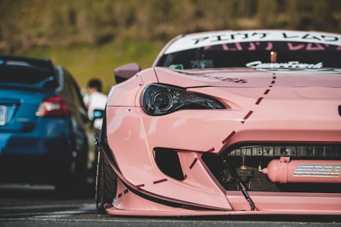 A close-up of a modified pink sports car with visible decals and custom bodywork. The focus is on the front end, showing intricate details such as a unique headlight design and body modifications. In the background, a blue car and a blurred figure are partially visible.