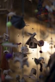 A wooden birdhouse is suspended in the center, adorned with a carved bird perched on the roof. Surrounding it are various hanging ornaments, including colorful felt balls, small animal figures, and other handmade decorations. The background features a warm, blurred light that adds a soft ambiance to the scene.