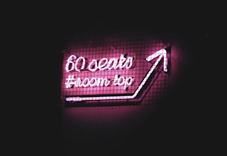 Neon sign with pink glowing text reads '60 seats #rooftop' and an arrow pointing upwards. The sign is set against a dark background with a grid overlay.