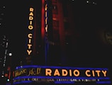 Neon signs with the text 'Radio City' and 'Music Hall' in bright orange and blue colors illuminate the facade of a building at night. Additional text below reads 'Freestyle & Old School Extravaganza February 9.' The scene includes vertical and horizontal signage with a dark surrounding environment.