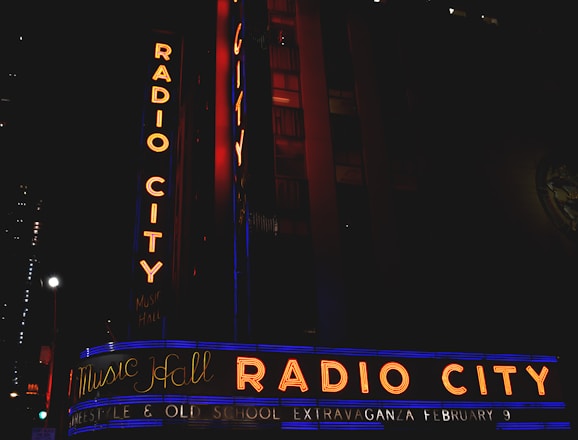 Neon signs with the text 'Radio City' and 'Music Hall' in bright orange and blue colors illuminate the facade of a building at night. Additional text below reads 'Freestyle & Old School Extravaganza February 9.' The scene includes vertical and horizontal signage with a dark surrounding environment.