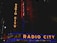 Neon signs with the text 'Radio City' and 'Music Hall' in bright orange and blue colors illuminate the facade of a building at night. Additional text below reads 'Freestyle & Old School Extravaganza February 9.' The scene includes vertical and horizontal signage with a dark surrounding environment.