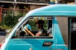 A person driving a teal-colored vehicle with the words 'Motorsport' and 'Special Edition' written on it. The vehicle is on a street with greenery and buildings in the background. The driver appears to be focused, holding the steering wheel with both hands.