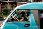 A person driving a teal-colored vehicle with the words 'Motorsport' and 'Special Edition' written on it. The vehicle is on a street with greenery and buildings in the background. The driver appears to be focused, holding the steering wheel with both hands.