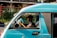 A person driving a teal-colored vehicle with the words 'Motorsport' and 'Special Edition' written on it. The vehicle is on a street with greenery and buildings in the background. The driver appears to be focused, holding the steering wheel with both hands.