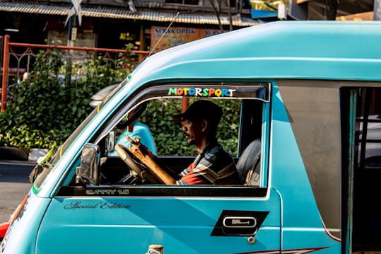 A person driving a teal-colored vehicle with the words 'Motorsport' and 'Special Edition' written on it. The vehicle is on a street with greenery and buildings in the background. The driver appears to be focused, holding the steering wheel with both hands.