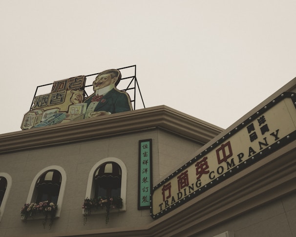A vintage-style building with a sign on the rooftop featuring an illustration of a well-dressed man holding products. The facade includes arched windows adorned with decorative flowers. Additional signs with Chinese characters and the words 'TRADING COMPANY' give the scene a historic or retro commercial appearance.
