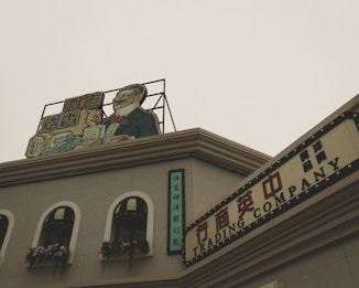 A vintage-style building with a sign on the rooftop featuring an illustration of a well-dressed man holding products. The facade includes arched windows adorned with decorative flowers. Additional signs with Chinese characters and the words 'TRADING COMPANY' give the scene a historic or retro commercial appearance.