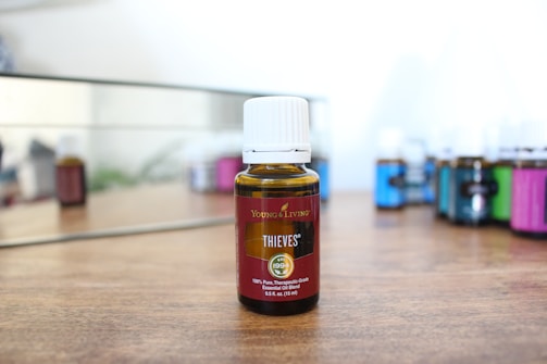 A bottle of essential oil labeled 'Thieves' from Young Living is placed on a wooden surface. The bottle has a white cap and a brown body. In the background, several other bottles of essential oil in various colors are slightly out of focus, reflecting on a mirror.
