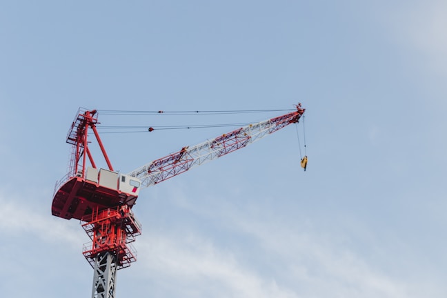 A large red and white construction crane extends its arm high into the sky, equipped with a hoisting mechanism. The crane is seen against a clear blue sky with minimal cloud cover.