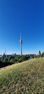 A tall communication tower rises prominently against a clear blue sky, surrounded by lush green trees and grass in the foreground. In the distance, modern architectural structures are visible, including a stadium-like construction and several buildings. The scene conveys a balance between nature and urban development.