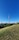 A tall communication tower rises prominently against a clear blue sky, surrounded by lush green trees and grass in the foreground. In the distance, modern architectural structures are visible, including a stadium-like construction and several buildings. The scene conveys a balance between nature and urban development.