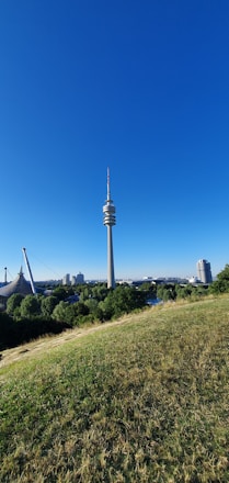 A tall communication tower rises prominently against a clear blue sky, surrounded by lush green trees and grass in the foreground. In the distance, modern architectural structures are visible, including a stadium-like construction and several buildings. The scene conveys a balance between nature and urban development.