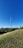 A tall communication tower rises prominently against a clear blue sky, surrounded by lush green trees and grass in the foreground. In the distance, modern architectural structures are visible, including a stadium-like construction and several buildings. The scene conveys a balance between nature and urban development.