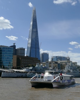 A riverboat with the Union Jack flag sails on a river in an urban area. In the background, a tall, modern glass skyscraper stands prominently among other buildings and old ships in a dock. The sky is clear with a few clouds.
