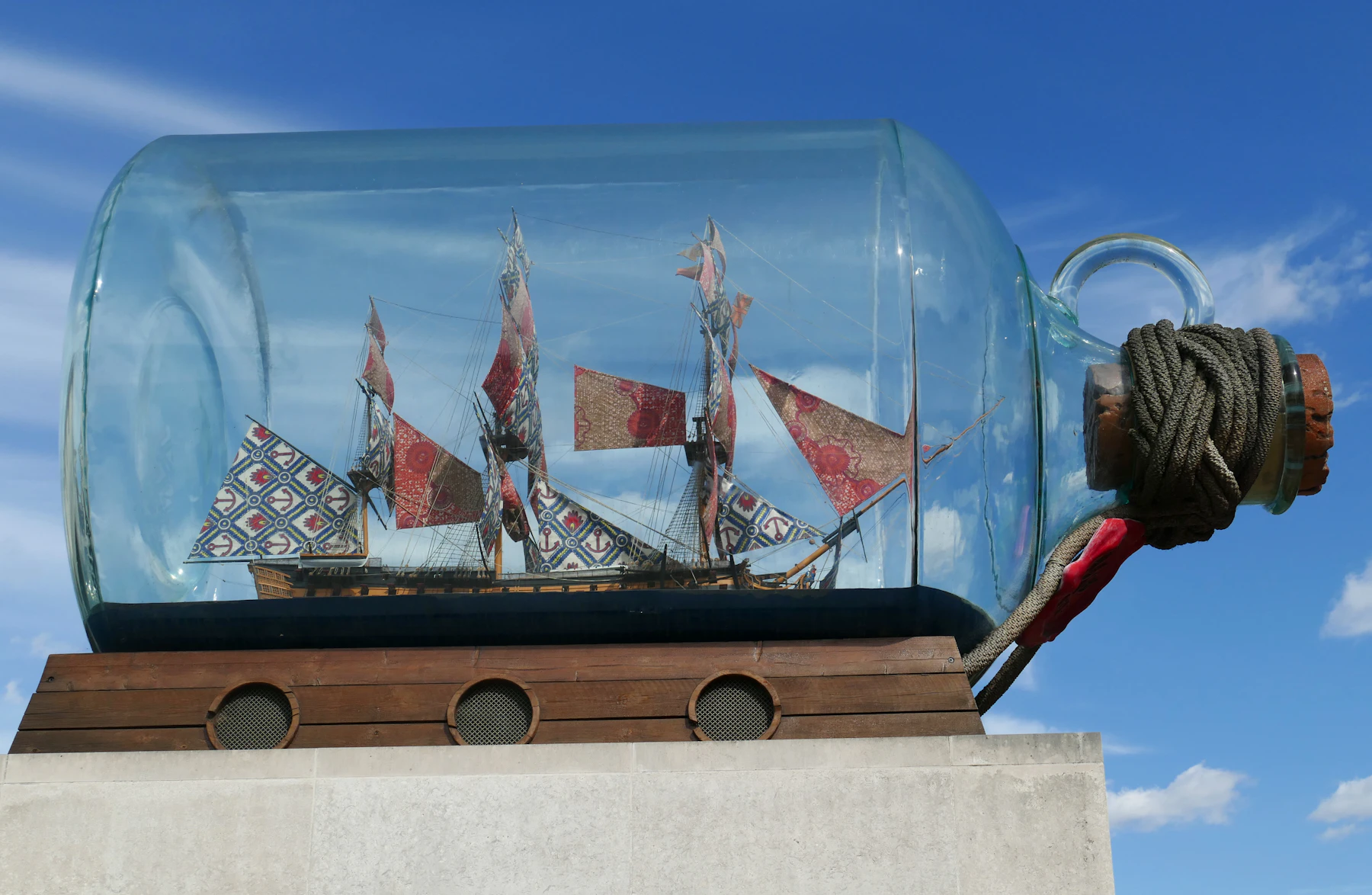 A large ship in a bottle with patterned sails and a rope-wrapped cork, on a plinth against a blue sky