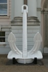 A large white anchor is displayed in front of a neoclassical building with columns and arched windows. The anchor is placed on a black platform and is positioned centrally, standing upright.