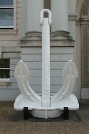 A large white anchor is displayed in front of a neoclassical building with columns and arched windows. The anchor is placed on a black platform and is positioned centrally, standing upright.