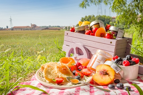 A picnic scene in a field with a wooden crate full of jars and fresh fruits like strawberries, peaches, and berries displayed on a checkered tablecloth. A jar of fruit preserve is open, with a spoon resting nearby. The background shows a lush green field, trees, and a few buildings in the distance under a clear blue sky.
