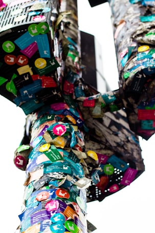 A pole is covered in a multitude of colorful stickers in various shapes and sizes. The stickers are layered on top of one another, creating a dense collage that includes arrows, logos, and text. The background is slightly out of focus, drawing attention to the vibrant display on the pole.