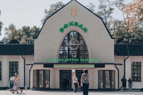 A beige building with dark roofing and large, arch-shaped windows is featured prominently. Above the entrance, vibrant green text is displayed. Several people are walking and standing around the entrance, with trees visible in the background under a clear sky.