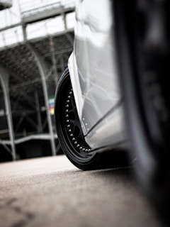 A close-up of a car's wheel and fender, showcasing the glossy black rim with metallic studs. The background features a blurred view of a metallic structure, possibly a grandstand or building, giving an industrial feel.