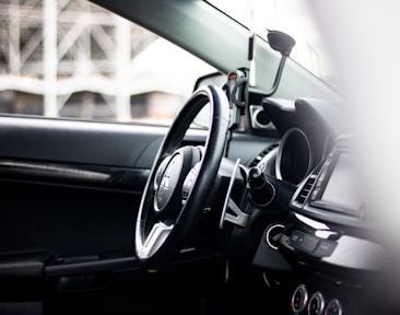 The interior of a car, focusing on the steering wheel and dashboard area with visible controls and gauges. A phone holder is mounted on the dashboard, and the overall view suggests a modern and clean vehicle interior.