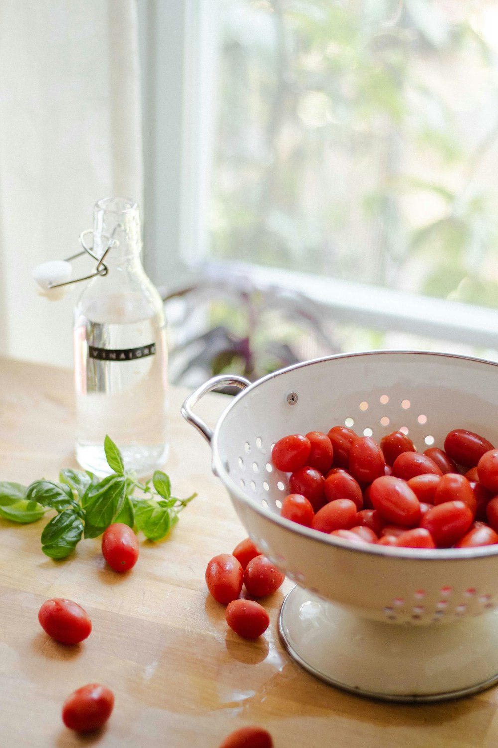 red tomatoes in round white strainer near green mint and glass bottle on table