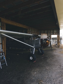 A vintage aircraft is parked in a dimly lit hangar with exposed wooden beams. The plane's nose and propeller are visible, while a metal ladder leans against the wall on the left.