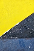 A close-up view of a wall with a textured surface, featuring a bright yellow section on the top and a rugged, dark blue area on the bottom. The paint appears cracked and peeling at the boundary where the two colors meet.