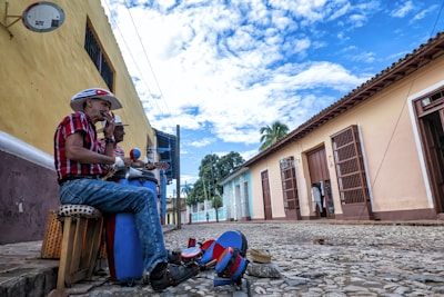 Two men wearing cowboy hats and plaid shirts are seated on stools along a cobblestone street, playing guitars. They are surrounded by colorful buildings with barred windows and doors, casting a lively and vibrant atmosphere. The sky above is partly cloudy, and the street is lined with traditional architecture, including a visible satellite dish.