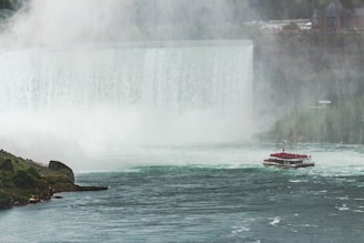 A large waterfall cascades down a steep cliff, creating a misty atmosphere. In the foreground, a boat filled with tourists in red ponchos navigates the turbulent waters near the waterfall. Lush greenery borders the waterway, and several buildings are visible in the distance, adding an element of civilization to the natural beauty.