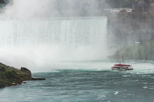 A large waterfall cascades down a steep cliff, creating a misty atmosphere. In the foreground, a boat filled with tourists in red ponchos navigates the turbulent waters near the waterfall. Lush greenery borders the waterway, and several buildings are visible in the distance, adding an element of civilization to the natural beauty.