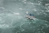 A boat filled with people wearing blue raincoats navigates through choppy waters. The vessel, named 'Maid of the...', appears to be a tour boat, possibly approaching a waterfall or turbulent water area. The surrounding water is turbulent and foamy, creating a sense of movement and energy.