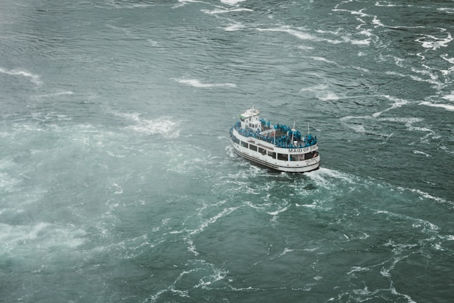 A boat filled with people wearing blue raincoats navigates through choppy waters. The vessel, named 'Maid of the...', appears to be a tour boat, possibly approaching a waterfall or turbulent water area. The surrounding water is turbulent and foamy, creating a sense of movement and energy.