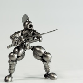 A small robot figure made from various metal components including screws, bolts, and nuts, posed in a dynamic, crouching stance as if holding a sword. The figure has a humanoid shape with detailed features such as a helmet-like head and articulated limbs, giving it an industrial, mechanical appearance.