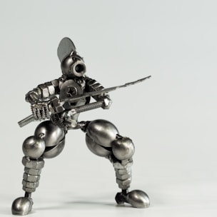 A small robot figure made from various metal components including screws, bolts, and nuts, posed in a dynamic, crouching stance as if holding a sword. The figure has a humanoid shape with detailed features such as a helmet-like head and articulated limbs, giving it an industrial, mechanical appearance.