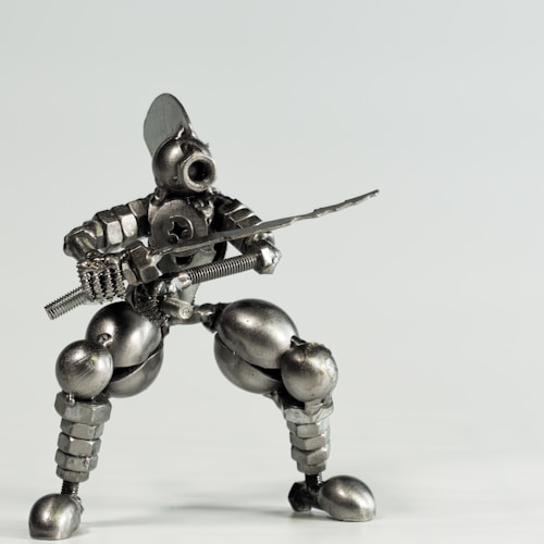 A small robot figure made from various metal components including screws, bolts, and nuts, posed in a dynamic, crouching stance as if holding a sword. The figure has a humanoid shape with detailed features such as a helmet-like head and articulated limbs, giving it an industrial, mechanical appearance.
