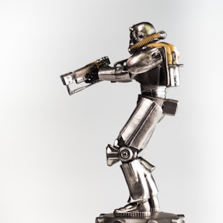 A metallic robot figurine is prominently displayed against a plain background. The robot is equipped with a helmet and a breathing apparatus attached to a backpack. Its body features detailed mechanical joints and armor plating, and it holds a futuristic rifle in its hands.