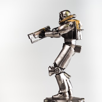 A metallic robot figurine is prominently displayed against a plain background. The robot is equipped with a helmet and a breathing apparatus attached to a backpack. Its body features detailed mechanical joints and armor plating, and it holds a futuristic rifle in its hands.