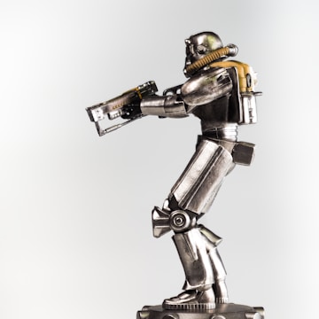 A metallic robot figurine is prominently displayed against a plain background. The robot is equipped with a helmet and a breathing apparatus attached to a backpack. Its body features detailed mechanical joints and armor plating, and it holds a futuristic rifle in its hands.