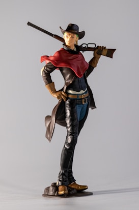 A figurine of a person dressed in a Western-style outfit, including a cowboy hat, red scarf, leather jacket, and holding a rifle. The figure stands confidently with one hand on the hip and the other holding the rifle behind the shoulder. The clothing includes black pants, a blue shirt, and brown gloves and boots. It is positioned on a small base, resembling rocky terrain.