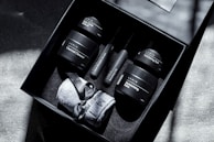 A black gift box contains an assortment of skincare products including a charcoal cleanser, dark circle defense, anti-wrinkle serum, recovery oil, moisturizing balm, and exfoliating rub. The box also includes a pair of grey socks tied with a ribbon. The arrangement is neatly placed on a textured surface with soft shadows.
