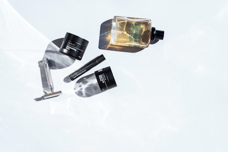 Several grooming products are artistically arranged on a white surface with subtle water ripple reflections. The items include a metal razor, a small tub labeled moisturizing balm, a cylindrical anti-wrinkle serum container, and a bottle of cologne. The lighting highlights the products and casts distinct shadows, creating a minimalist and clean aesthetic.