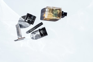Several grooming products are artistically arranged on a white surface with subtle water ripple reflections. The items include a metal razor, a small tub labeled moisturizing balm, a cylindrical anti-wrinkle serum container, and a bottle of cologne. The lighting highlights the products and casts distinct shadows, creating a minimalist and clean aesthetic.