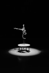 A person is performing an impressive handstand on a tall stand atop a circular platform, lit by a spotlight in an otherwise dark space. The individual appears to be shirtless and muscular, showcasing strength and balance. Shadows cast on the ground create a dramatic effect.