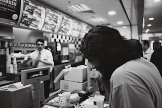 A black and white photograph captured inside a fast-food restaurant. A customer with long hair is leaning over the counter, which is cluttered with cups and food items. In the background, an employee dressed in a uniform is visible, seemingly interacting with another customer. The menu above displays various food options, and the digital display on the register reads, 'AFIYET OLSUN.' The restaurant is moderately busy, with other patrons in the background.
