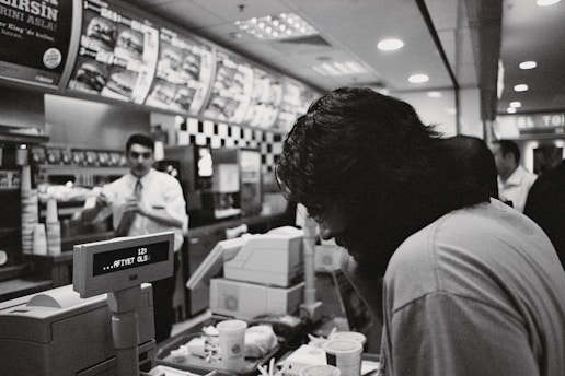 A black and white photograph captured inside a fast-food restaurant. A customer with long hair is leaning over the counter, which is cluttered with cups and food items. In the background, an employee dressed in a uniform is visible, seemingly interacting with another customer. The menu above displays various food options, and the digital display on the register reads, 'AFIYET OLSUN.' The restaurant is moderately busy, with other patrons in the background.